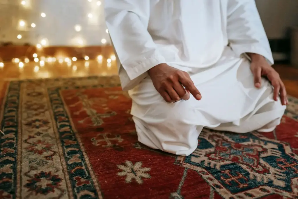 A man in a white robe kneeling on a red prayer rug