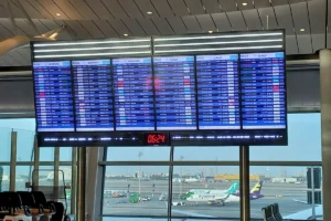Flight information screens in the terminal of King Abdulaziz International Airport, Jeddah, with aircraft on the apron behind