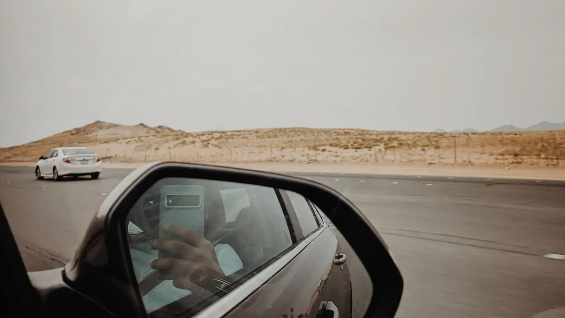Desert highway and sand dunes seen in a car side mirror in Saudi Arabia; the exact road is not stated