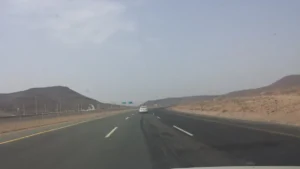 Divided highway toward Makkah beside Al Jumum, with rocky hills and a green direction sign ahead