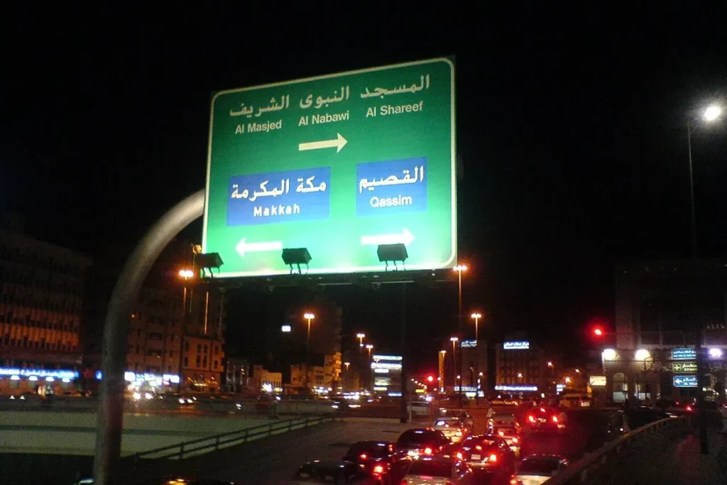 Green road sign in Madinah at night pointing to Al Masjid Al Nabawi, Makkah and Qassim, over evening traffic