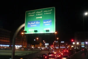 Green road sign in Madinah at night pointing to Al Masjid Al Nabawi, Makkah and Qassim, over evening traffic