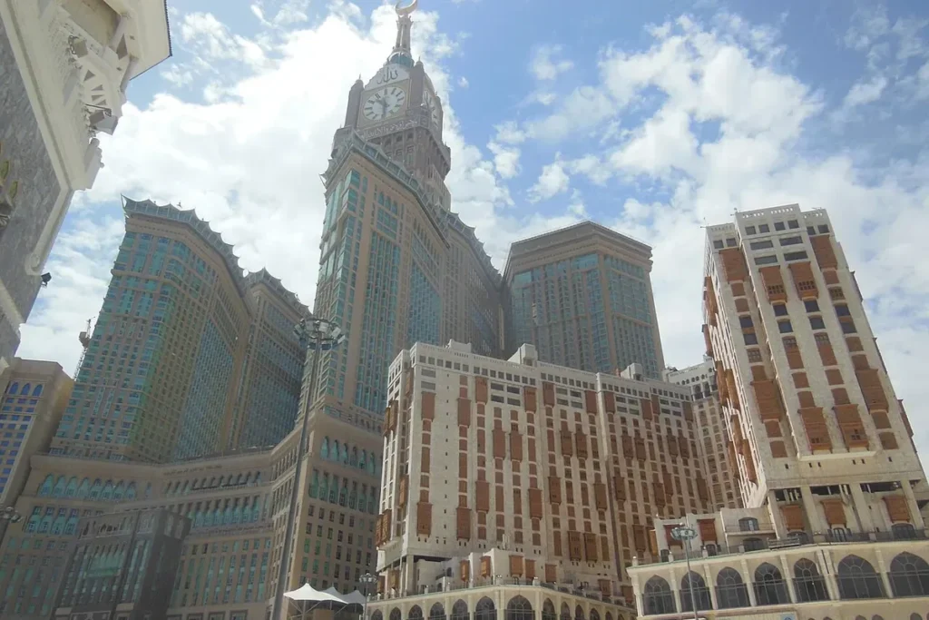 Abraj Al Bait clock tower and neighbouring hotel towers from street level under a cloudy sky