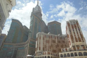 Abraj Al Bait clock tower and neighbouring hotel towers from street level under a cloudy sky