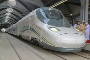 Haramain High Speed Railway Talgo 350 train, white with green and gold stripes, at a station platform; the station is not named by the source