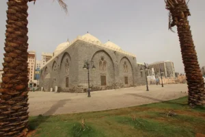 Masjid al-Ghamama framed by palm trunks across the open plaza